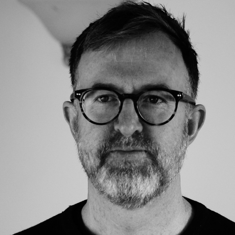 Dr Illan Wall stands in front of a white wall wearing a black t-shirt in this black and white image.