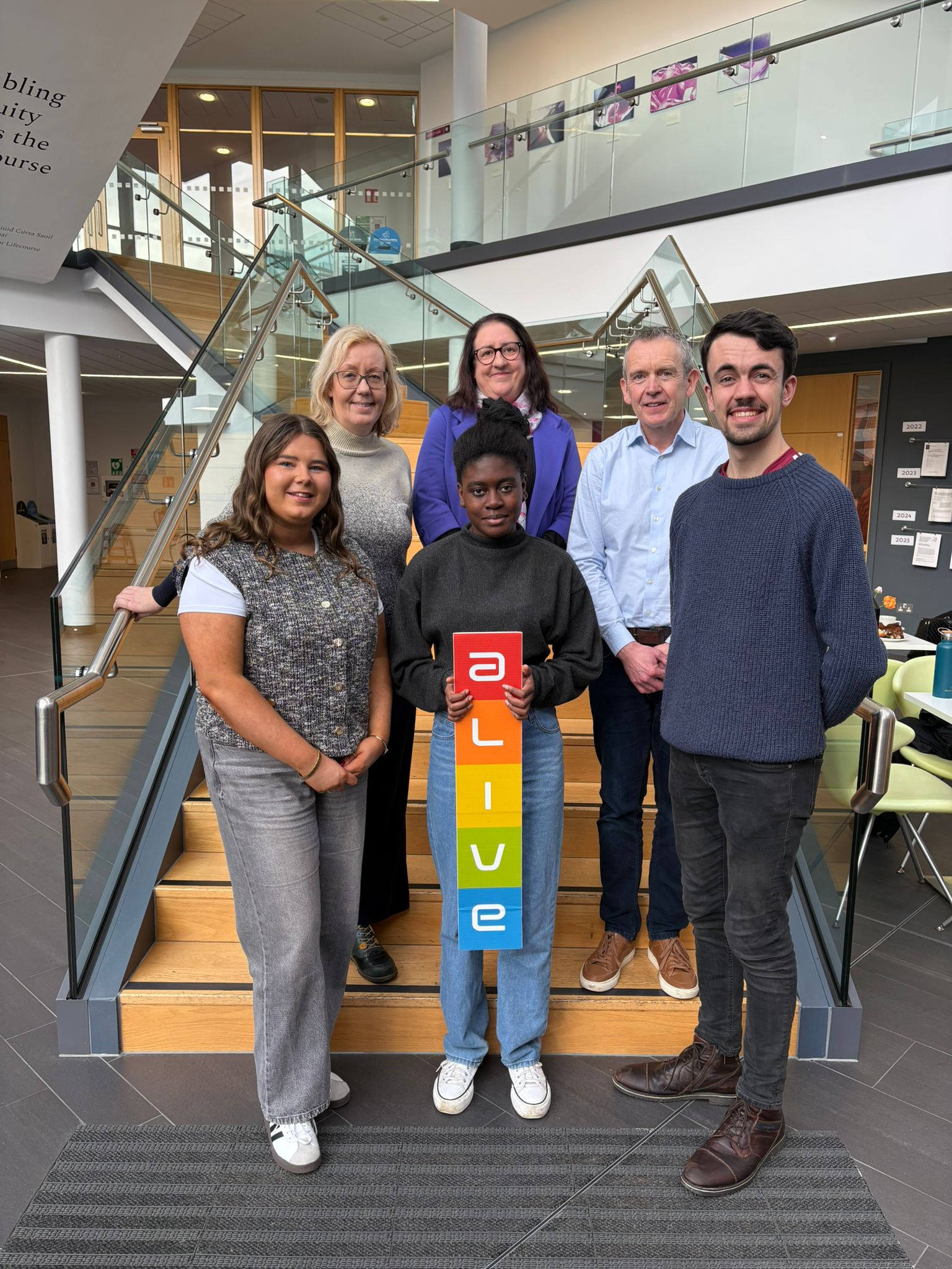 6 people standing in front of stairs, one of them holds the ALIVE sign.